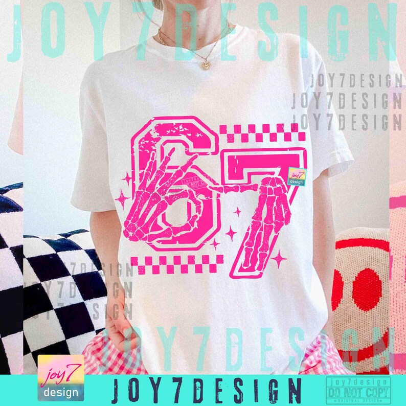 Peut inclure: T-shirt blanc avec un motif rose du chiffre 67 orn&eacute; de mains de squelette, d'un motif &agrave; carreaux et d'&eacute;toiles. Le texte "JOY7DESIGN" est &eacute;galement pr&eacute;sent. Un t-shirt d&eacute;contract&eacute; et graphique.