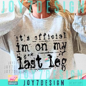 May include: A light gray sweatshirt with the text "it's official im on my last leg" in black. The sweatshirt has a crew neck and long sleeves. The design is a distressed, vintage style.