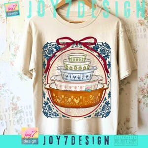 May include: A beige t-shirt featuring a graphic of a stack of vintage casserole dishes in various colors and patterns. The design is framed by a red ribbon and a blue and white floral border. The text "JOY7DESIGN" is visible.