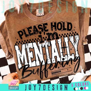 May include: A brown Comfort Colors t-shirt with the text "PLEASE HOLD I'M MENTALLY Buffering" in black and white. The shirt has a distressed look and a checkered pattern. The design includes a loading bar graphic.