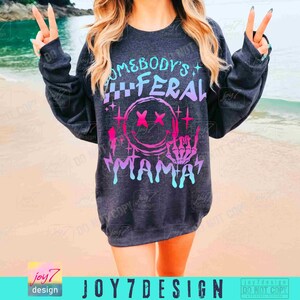 K&ouml;nnte beinhalten: Ein dunkelgraues Sweatshirt mit einem pinkfarbenen, blauen und wei&szlig;en Grafikdesign, auf dem "Somebody's Feral Mama" mit einem Smiley und einer Skeletthand, die ein Rock'n'Roll-Zeichen macht, steht.