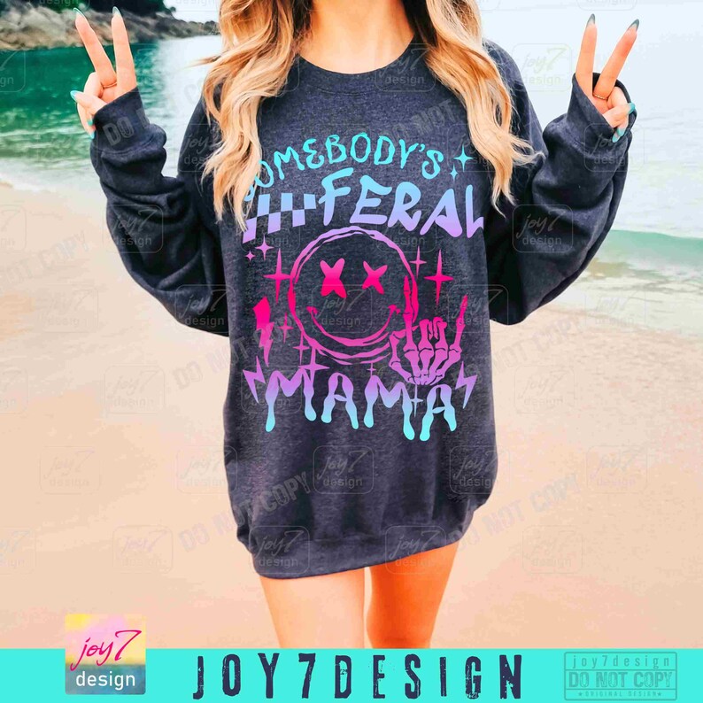 K&ouml;nnte beinhalten: Ein dunkelgraues Sweatshirt mit einem pinkfarbenen, blauen und wei&szlig;en Grafikdesign, auf dem "Somebody's Feral Mama" mit einem Smiley und einer Skeletthand, die ein Rock'n'Roll-Zeichen macht, steht.