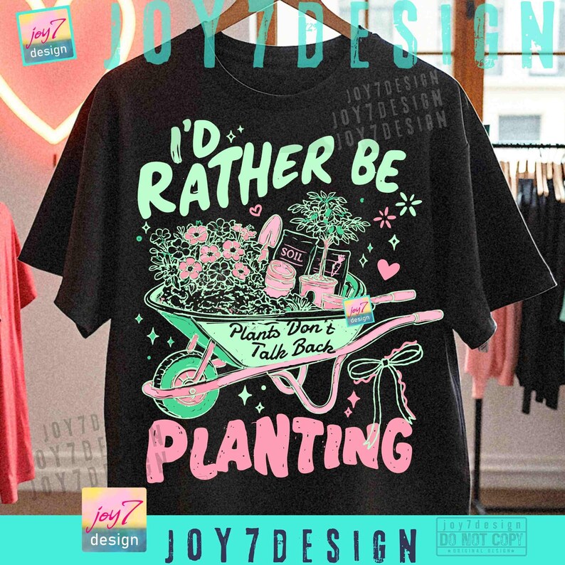 May include: Black t-shirt with a gardening-themed graphic. The design features a wheelbarrow filled with flowers, a small tree, and the text "I'd Rather Be Planting." The text "Plants Don't Talk Back" is also included.