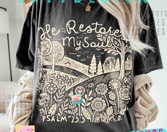 Op de afbeelding: Zwart t-shirt met een crèmekleurige print. De print toont de tekst "He Restore My Soul" boven een landschapsillustratie met bomen, heuvels en bloemen. De tekst "PSALM 23:3" staat eronder.