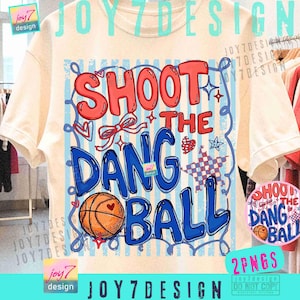 May include: Cream-colored t-shirt with a basketball-themed graphic. The design features the text "SHOOT THE DANG BALL" in red and blue, along with a basketball, stars, and a bow. The shirt has short sleeves.