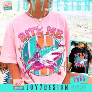 May include: A light pink t-shirt featuring a graphic of a shark in shades of blue, pink, and orange. The text BITE ME is printed in a bold, retro-style font above the shark. The design includes star and lightning bolt accents within a circular frame.