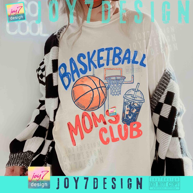 May include: Cream-colored t-shirt with a basketball graphic. The design includes a basketball, hoop, and a drink, with the words "BASKETBALL MOMS CLUB" in blue and red. The person is wearing a black and white checkered cardigan.