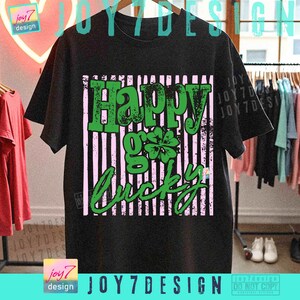 Może przedstawiać: Czarny t-shirt z zielono-r&oacute;żową grafiką. Grafika głosi "Happy Go Lucky" z czterolistną koniczyną. Koszulka wisi na drewnianym wieszaku z innymi ubraniami. Widoczna jest nazwa marki "joy7 design".