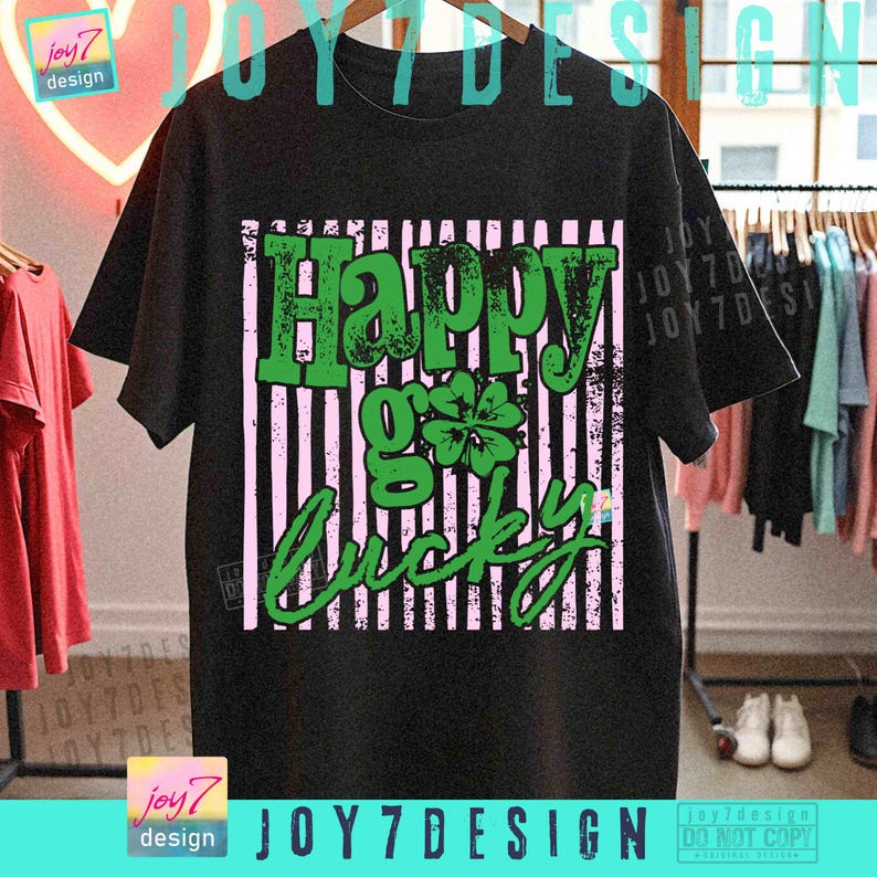 Może przedstawiać: Czarny t-shirt z zielono-r&oacute;żową grafiką. Grafika głosi "Happy Go Lucky" z czterolistną koniczyną. Koszulka wisi na drewnianym wieszaku z innymi ubraniami. Widoczna jest nazwa marki "joy7 design".