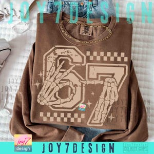 Peut inclure: Un sweat-shirt marron avec un motif vieilli repr&eacute;sentant le chiffre "67" et des mains de squelette. Le motif comprend un motif &agrave; carreaux et des &eacute;toiles. Le texte "JOY7DESIGN" est visible en bas.