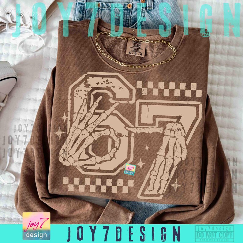 Peut inclure: Un sweat-shirt marron avec un motif vieilli repr&eacute;sentant le chiffre "67" et des mains de squelette. Le motif comprend un motif &agrave; carreaux et des &eacute;toiles. Le texte "JOY7DESIGN" est visible en bas.