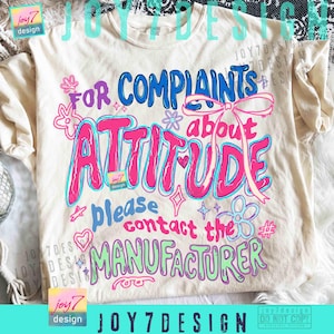 May include: Cream-colored t-shirt with the text "FOR COMPLAINTS ABOUT ATTITUDE please contact the MANUFACTURER" in blue, pink, and green. The design includes floral and heart accents.
