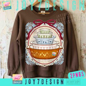 May include: Brown sweatshirt featuring a graphic of vintage casserole dishes in a decorative frame. The design includes a red ribbon and a blue and white lace-like border. The text "joy7 design" is visible.
