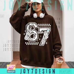 Peut inclure: Un sweat-shirt marron avec un motif blanc du chiffre 67, repr&eacute;sentant des mains de squelette. Le motif comprend un motif &agrave; carreaux et des &eacute;toiles. Le texte "JOY7DESIGN" est visible en bas.
