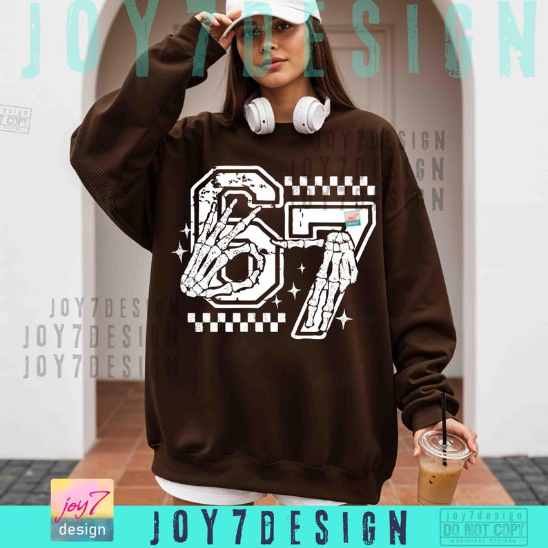 Peut inclure: Un sweat-shirt marron avec un motif blanc du chiffre 67, repr&eacute;sentant des mains de squelette. Le motif comprend un motif &agrave; carreaux et des &eacute;toiles. Le texte "JOY7DESIGN" est visible en bas.