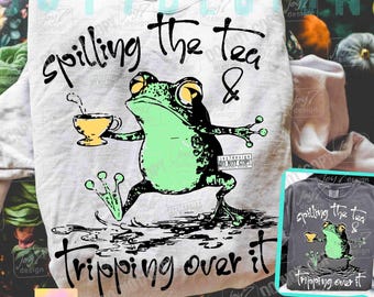 May include: A light gray sweatshirt with a graphic of a green frog holding a teacup, with the text "spilling the tea & tripping over it." The design includes a smaller image of the design on a dark gray shirt.
