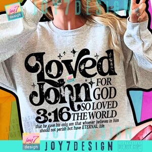 Op de afbeelding: Een lichtgrijs sweatshirt met de tekst "Loved For John 3:16 For God So Loved The World" in een zwarte, verweerde lettertype. Het sweatshirt bevat ook het vers "that he gave his only son that whoever believes in him should not perish but have ETERNAL life."