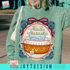 May include: A teal sweatshirt featuring a graphic of vintage Pyrex nesting bowls in various sizes and patterns. The design includes a red ribbon, a blue floral border, and the text "JOY7 DESIGN".