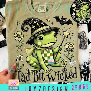 Op de afbeelding: Olijfgroen t-shirt met een cartoon kikker met een heksenhoed en een sterrenstaf. De kikker is groen met een dambordpatroon. De tekst "Tad Bit Wicked" is te zien. Extra afbeeldingen zijn bloemen en een vleermuis.