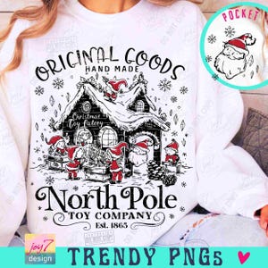May include: White sweatshirt featuring a black and red graphic of a Christmas toy factory with Santa Claus and elves. The text reads "Original Goods Hand Made" and "North Pole Toy Company Est. 1865". A pocket design shows Santa's face.