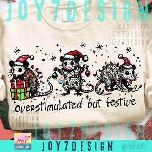 Puede incluir: Sudadera crema con tres zarig&uuml;eyas con gorros de Pap&aacute; Noel y luces navide&ntilde;as. Una zarig&uuml;eya sostiene un regalo. El texto "Overstimulated but festive" est&aacute; debajo. Ropa festiva para las fiestas.
