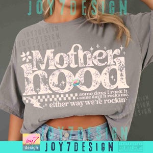 Peut inclure: Un t-shirt gris avec le mot "Motherhood" en grand, dans une police d&eacute;corative orn&eacute;e de motifs floraux et d'&eacute;toiles. La phrase "some days I rock it, some days it rocks me, either way we're rockin'" est imprim&eacute;e en dessous.