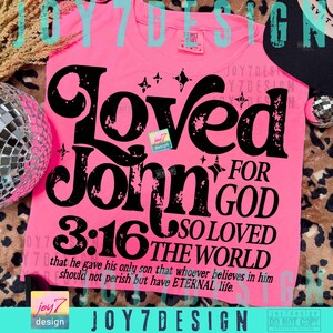 Op de afbeelding: Een felroze t-shirt met de tekst "Loved John 3:16" in grote, zwarte letters. De tekst bevat de zin "For God so loved the world" en een bijbelvers. Het shirt heeft een ronde hals.