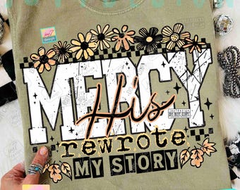May include: Olive green t-shirt with the words "MERCY His rewrote MY STORY" in a distressed, blocky font. The design includes floral and star accents. The shirt has a crew neck and short sleeves.