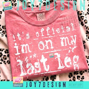 May include: A coral-colored t-shirt with the text "It's official, I'm on my last leg" in white, distressed lettering. The shirt has a crew neck and is made of a soft material. The design includes the brand name "JOY7DESIGN".