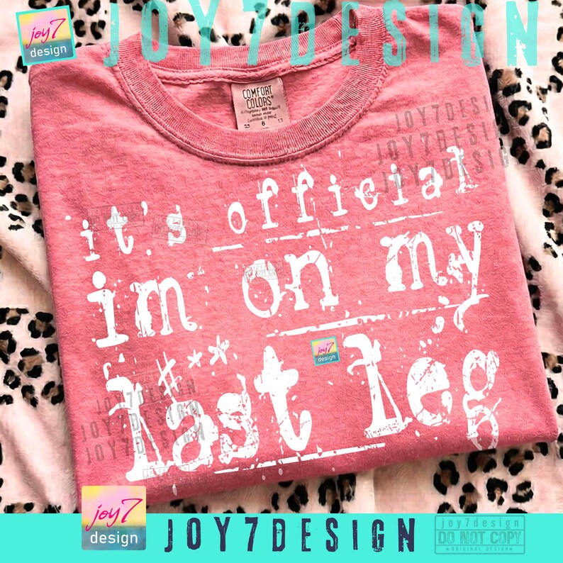 May include: A coral-colored t-shirt with the text "It's official, I'm on my last leg" in white, distressed lettering. The shirt has a crew neck and is made of a soft material. The design includes the brand name "JOY7DESIGN".