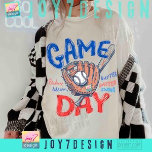May include: Cream-colored t-shirt with a baseball-themed graphic. The design features the words "GAME DAY" in blue and red, a baseball glove, a baseball, and a bat. The words "Baseball Season", "Batter, Batter, Swing" are also included.
