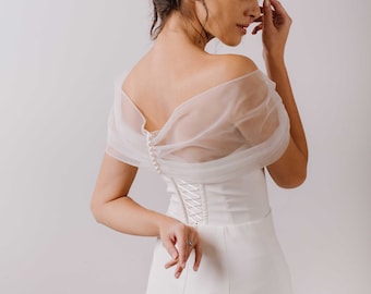 May include: A white off-the-shoulder wedding dress with a lace-up back and a sheer overlay. The dress has a fitted bodice and a flowing skirt.