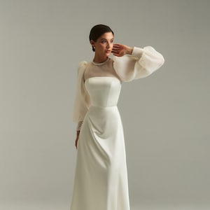 May include: A white satin wedding dress with a sheer, long-sleeved overlay and a high neckline. The dress has a fitted bodice and a flowing skirt.