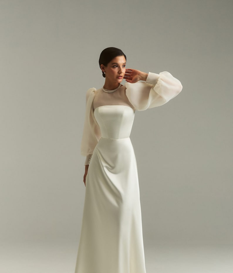 May include: A white satin wedding dress with a sheer, long-sleeved overlay and a high neckline. The dress has a fitted bodice and a flowing skirt.