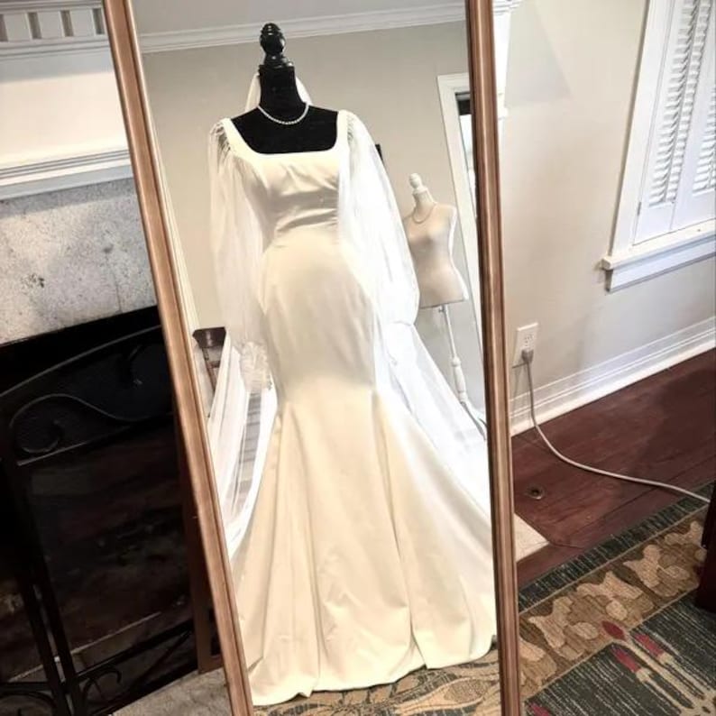 Pode incluir: Um vestido de noiva branco com um corpete justo e uma saia fluida. O vestido tem mangas compridas de tecido transparente e um decote quadrado. O vestido est&aacute; em um manequim em frente a um espelho.