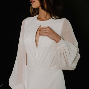 May include: A white long-sleeved dress with a keyhole neckline and a zip closure. The dress has a fitted bodice and a flowing skirt.