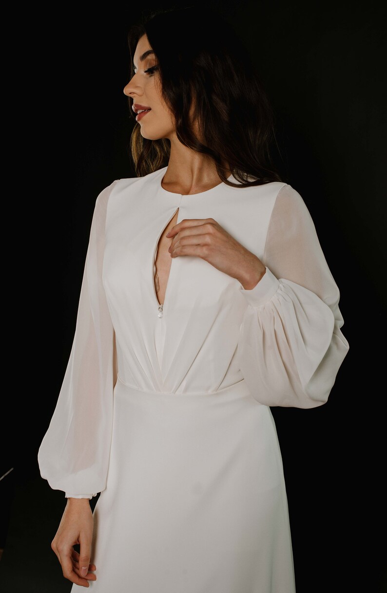 May include: A white long-sleeved dress with a keyhole neckline and a zip closure. The dress has a fitted bodice and a flowing skirt.