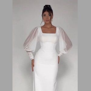 Pode incluir: Um vestido branco com decote quadrado e mangas compridas transparentes. O vestido tem um corpete justo e uma saia fluida.