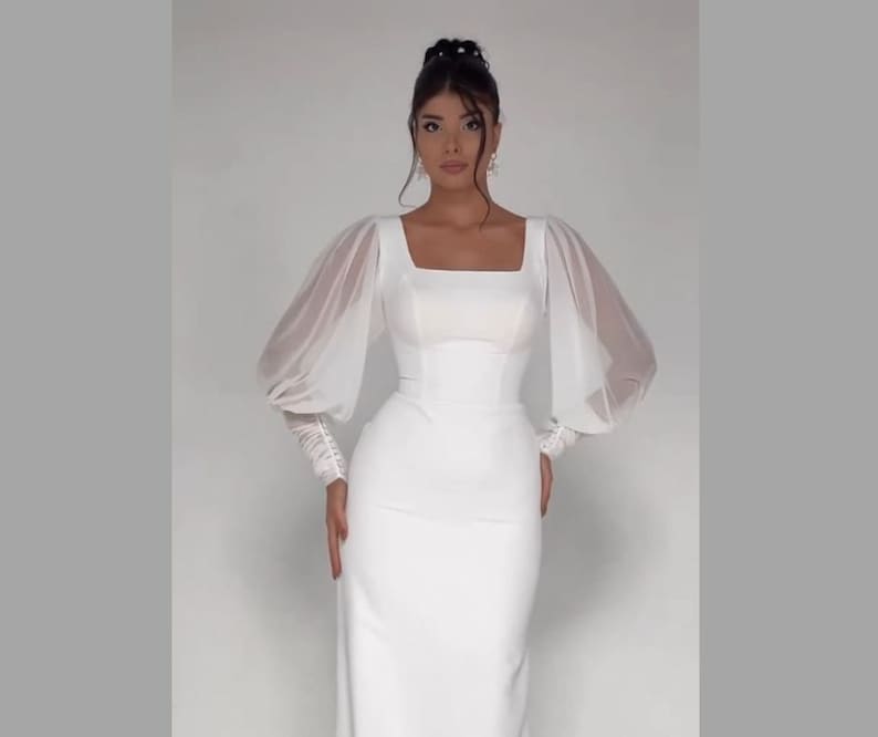 Pode incluir: Um vestido branco com decote quadrado e mangas compridas transparentes. O vestido tem um corpete justo e uma saia fluida.