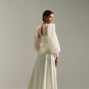 May include: A white satin wedding dress with a sheer, long-sleeved bodice and a fitted waist. The dress has a flowing skirt with a slit in the front.
