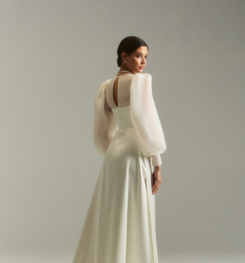May include: A white satin wedding dress with a sheer, long-sleeved bodice and a fitted waist. The dress has a flowing skirt with a slit in the front.