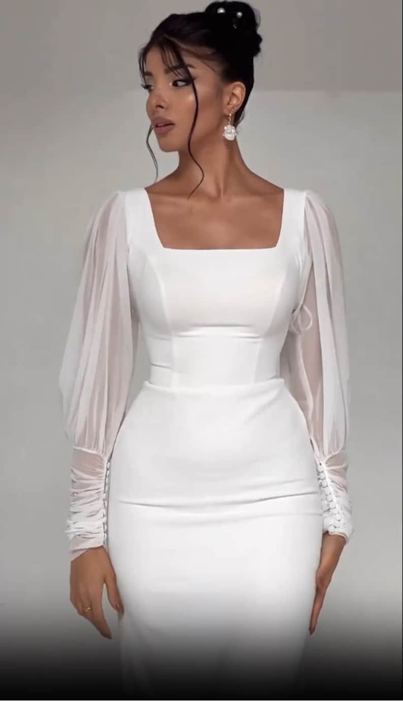 Pode incluir: Um vestido branco com mangas compridas e decote quadrado. As mangas s&atilde;o feitas de um tecido transparente e t&ecirc;m um detalhe franzido nos punhos.