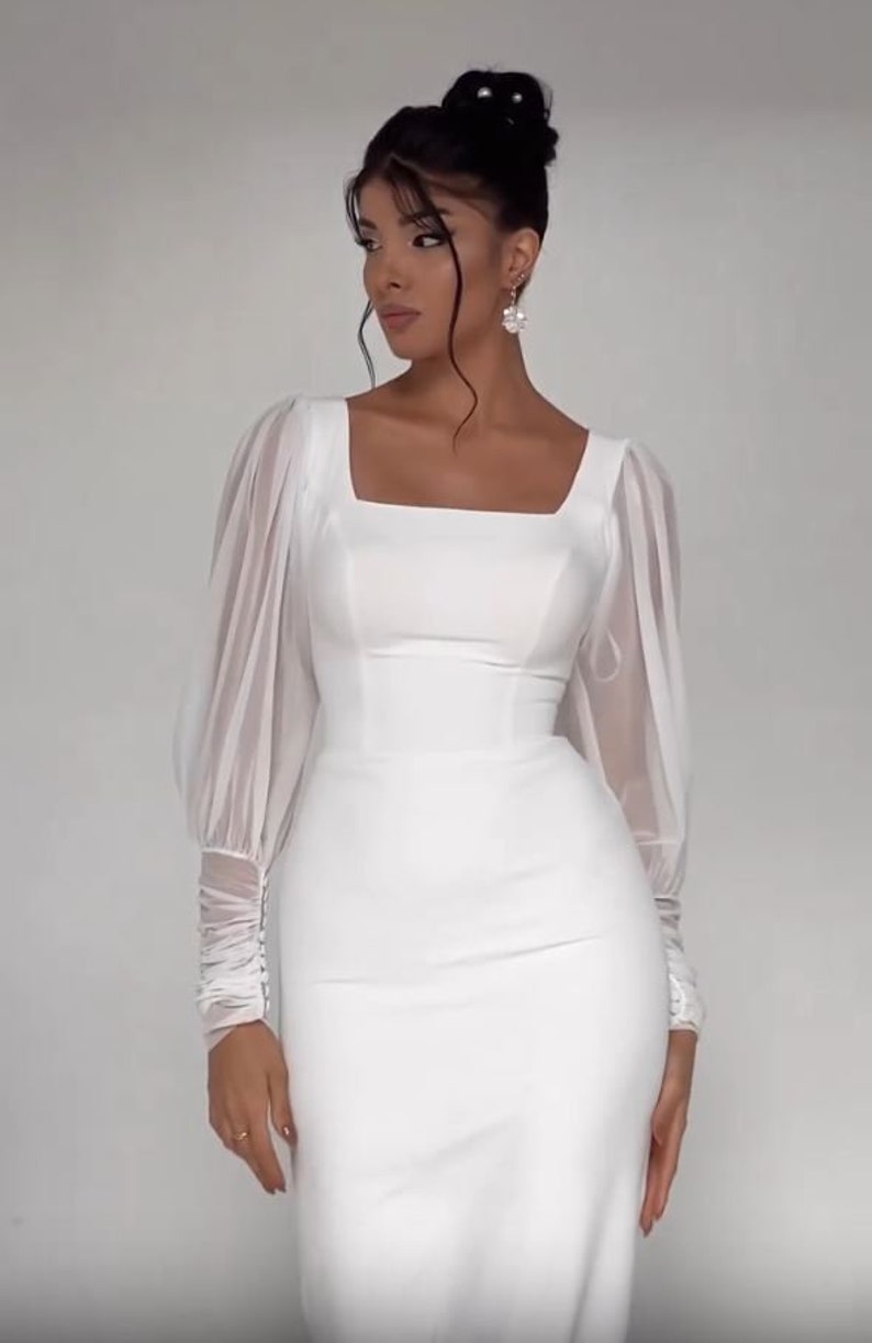 Pode incluir: Um vestido branco de manga comprida com decote quadrado e corpo justo. O vestido tem mangas bufantes transparentes com punhos franzidos.