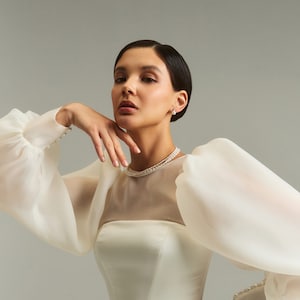 May include: A woman wearing a white wedding dress with a sheer bodice and large, puffy sleeves. The dress has a fitted bodice and a full skirt. She is wearing a pearl necklace and earrings.