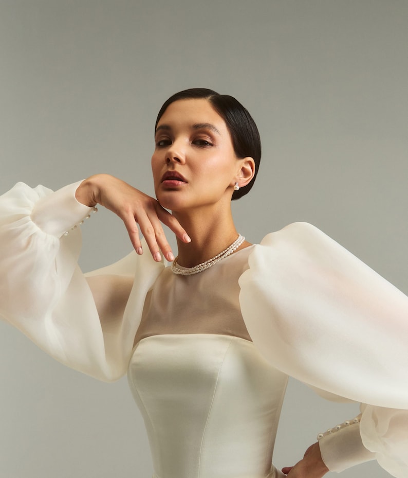May include: A woman wearing a white wedding dress with a sheer bodice and large, puffy sleeves. The dress has a fitted bodice and a full skirt. She is wearing a pearl necklace and earrings.
