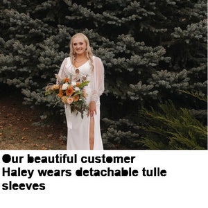 Pode incluir: Uma mulher com um vestido de noiva branco com uma fenda na saia e mangas de tule posa em frente a uma &aacute;rvore verde de folha perenne. Ela est&aacute; segurando um buqu&ecirc; de flores.