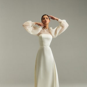 May include: A white wedding dress with a fitted bodice and a flowing skirt. The dress has sheer, puffed sleeves and a high neckline.