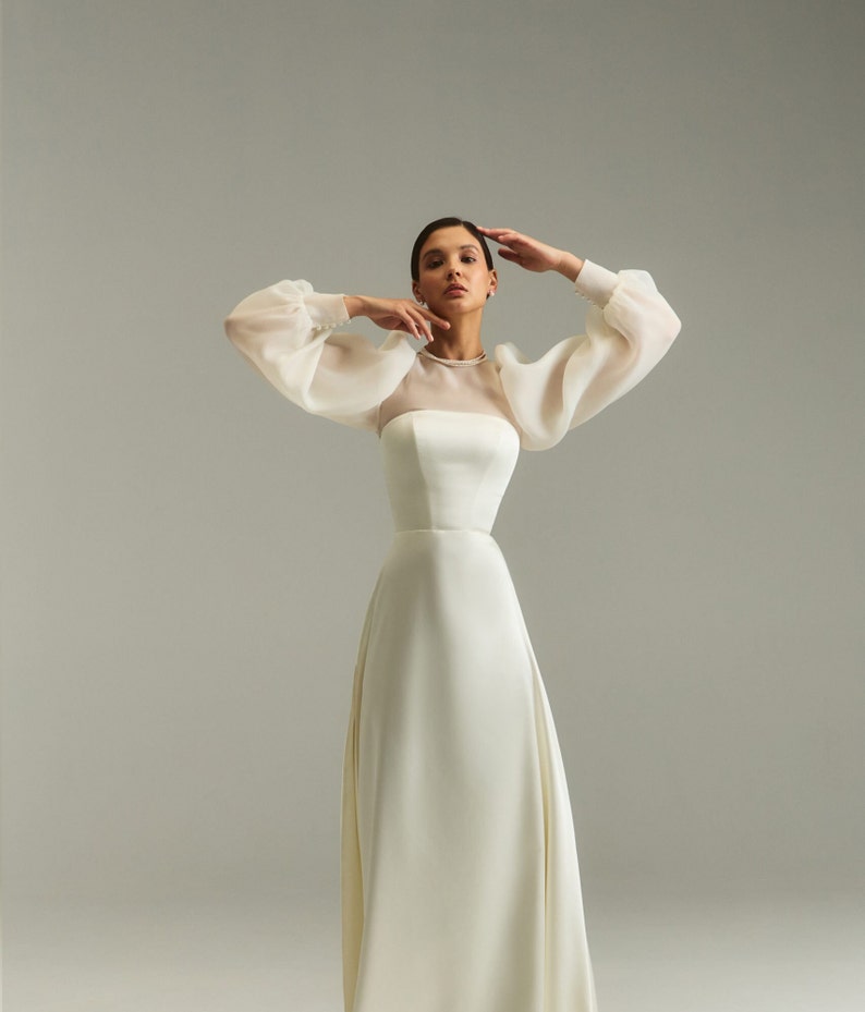 May include: A white wedding dress with a fitted bodice and a flowing skirt. The dress has sheer, puffed sleeves and a high neckline.