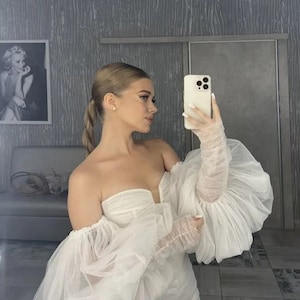 Könnte beinhalten: Eine Frau trägt ein weißes, schulterfreies Kleid mit großen, bauschigen Ärmeln. Das Kleid ist aus einem durchsichtigen Stoff und hat einen herzförmigen Ausschnitt. Die Frau hält ein weißes Smartphone in ihrer rechten Hand und schaut in die Kamera.