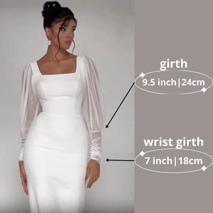 Pode incluir: Vestido branco com mangas compridas e corpo ajustado. As mangas s&atilde;o transparentes e franzidas no pulso. O vestido tem um decote quadrado. A imagem inclui as medidas da circunfer&ecirc;ncia do pulso (7 polegadas | 18 cm) e a circunfer&ecirc;ncia do peito (9,5 polegadas | 24 cm).